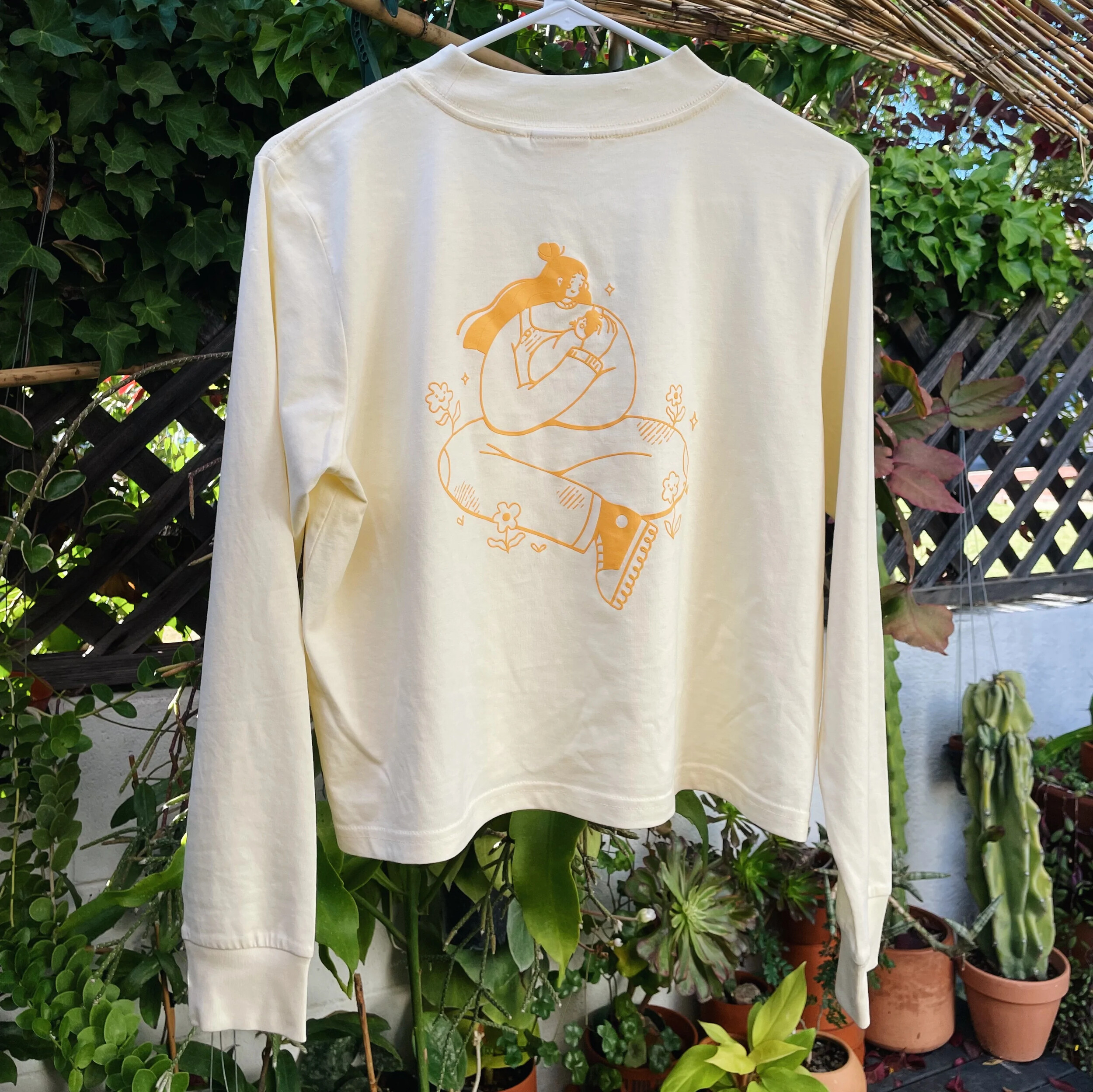 Photo of a cream long-sleeve shirt printed with a line-art mother and baby design, hanging among potted plants